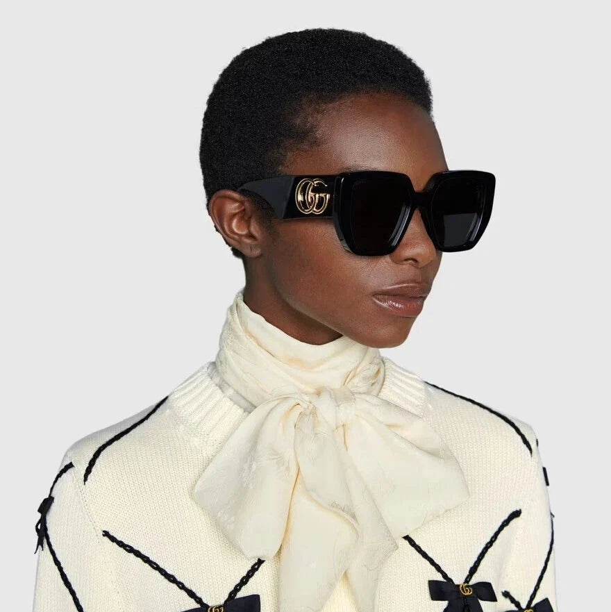 Woman wearing luxury black square sunglasses with gold logo and a cream neck scarf.